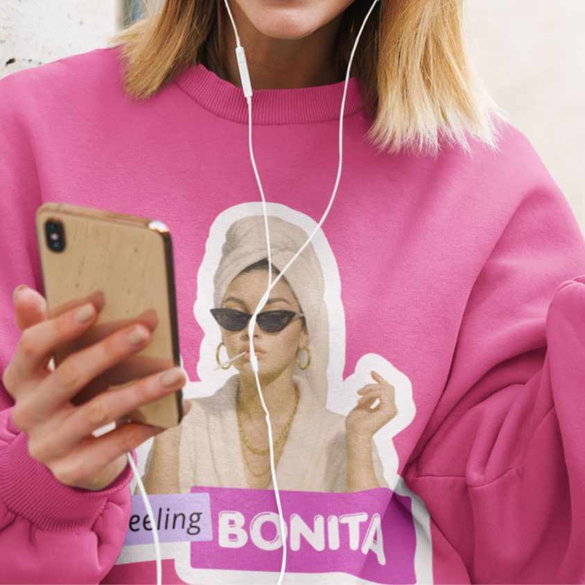 Woman in pink sweatshirt with 'feeling BONITA' design, using phone and earphones outdoors.
